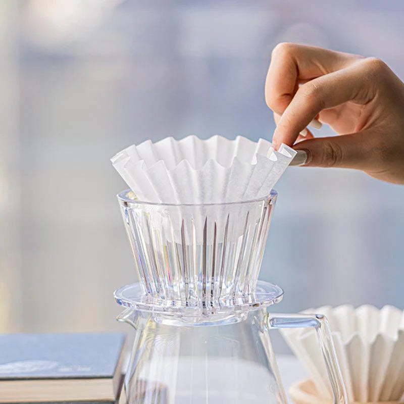 Hand placing a coffee filter paper into a clear glass coffee maker with a blurred background.
