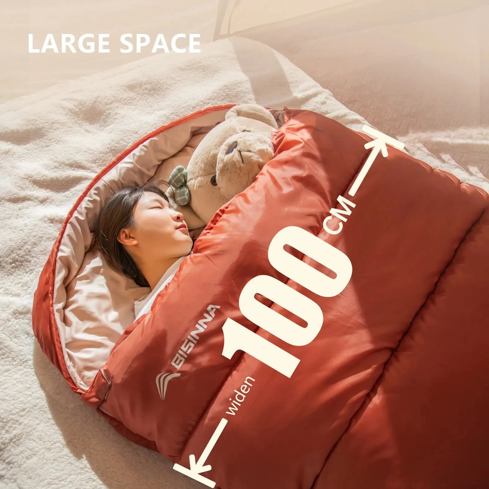 Person lying in a large BISNNA red sleeping bag with a teddy bear, labelled '100 cm', on a soft surface.