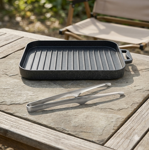A rectangular black non-stick grill pan with ridges and two side handles sits on a stone tabletop outdoors, with metal tongs placed in front and a folding chair blurred in the background.