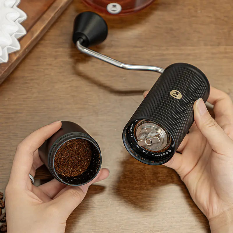 Person holding a TIMEMORE C5 ESP Pro manual coffee grinder with ground coffee on a wooden surface