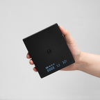 Hand holding a black TIMEMORE Black Mirror Mini coffee scales with a  digital display on a grey background.