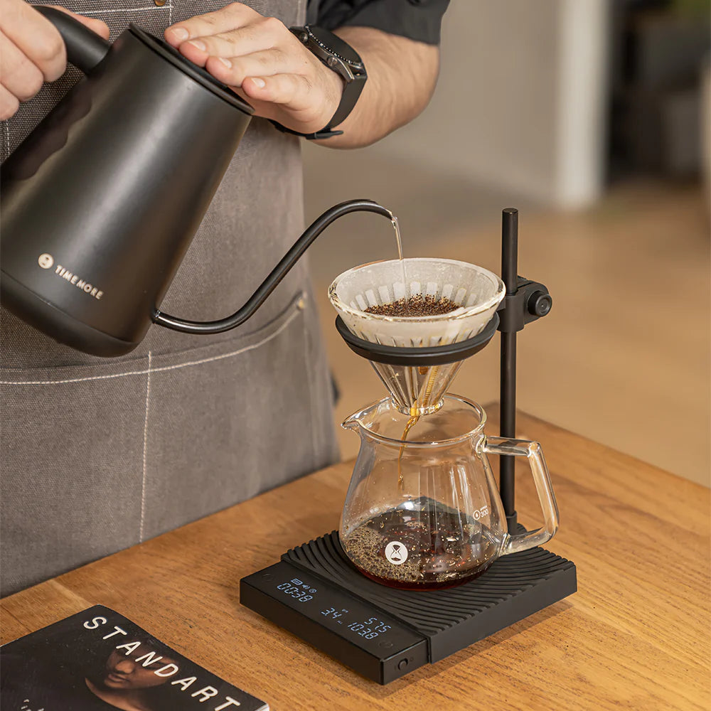 Person pouring coffee from a black kettle into a glass carafe with a coffee filter on a wooden surface.