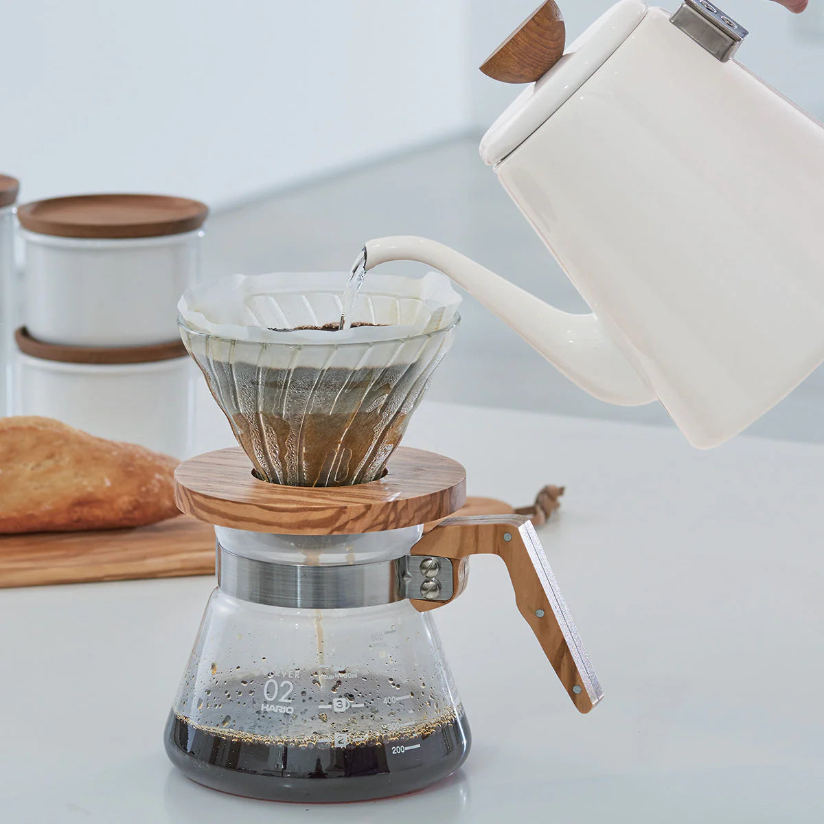 White kettle pouring water through a V60 filter into a glass coffee maker on a light surface.