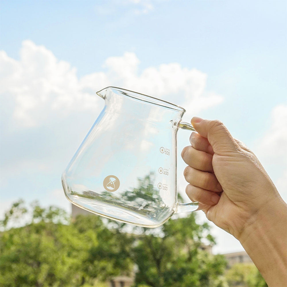 Hand holding a clear TIMEMORE Borosilicate Glass pitcher with a blurred outdoor background